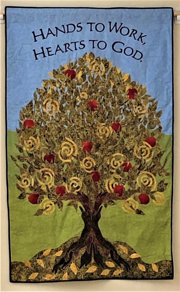 fabric art banner depicting the tree of life in greens and with red apples with the Celtic spiral in the tree leaveswith the Shaker proverb "hands to work, hearts to god"